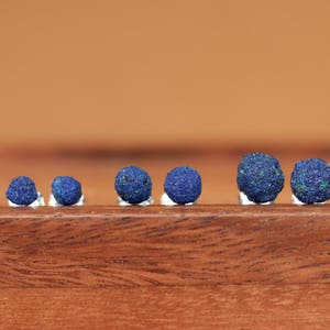 May include: A set of six small, round, blue gemstone earrings. The earrings are arranged in a row on a wooden surface.