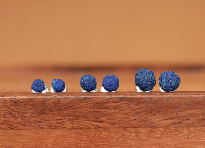 May include: A set of six small, round, blue gemstone earrings. The earrings are arranged in a row on a wooden surface.