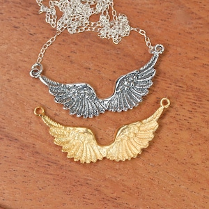 Puede incluir: Dos abalorios de alas de &aacute;ngel de plata, uno en un collar de cadena de plata y el otro en una cadena separada. Las alas de &aacute;ngel son detalladas y tienen un acabado texturizado. El collar est&aacute; sobre una superficie de madera.