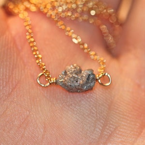 May include: A delicate gold chain necklace with a small, rough, grey diamond pendant.