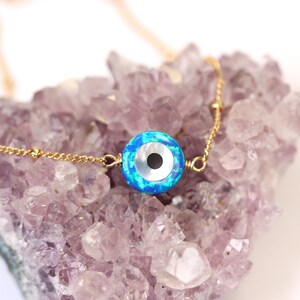 May include: A gold chain necklace with a blue and white opal evil eye charm. The charm is round and has a black pupil. The necklace is on a bed of purple amethyst crystals.