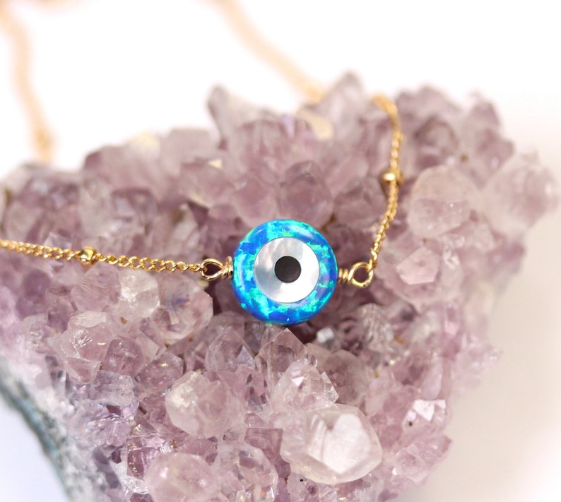 May include: A gold chain necklace with a blue and white opal evil eye charm. The charm is round and has a black pupil. The necklace is on a bed of purple amethyst crystals.
