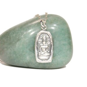 May include: Silver pendant necklace featuring a detailed image of a seated Buddha in a silver frame. The pendant is on a silver chain and is resting on a green stone.