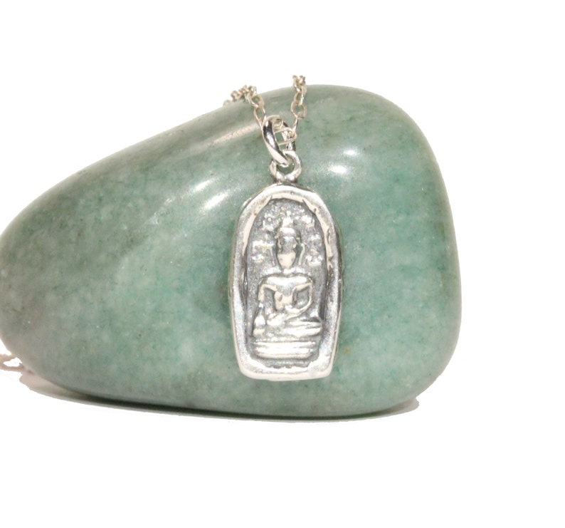 May include: Silver pendant necklace featuring a detailed image of a seated Buddha in a silver frame. The pendant is on a silver chain and is resting on a green stone.