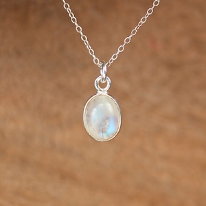 Puede incluir: Un collar de plata con un colgante ovalado de piedra lunar. La piedra lunar muestra una calidad lechosa e iridiscente con toques de azul y blanco. La delicada cadena de plata complementa el colgante, creando una joya sencilla y elegante.