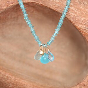 May include: A delicate necklace made with light blue gemstone beads and a charm of three smaller blue gemstones. The necklace is displayed on a wooden surface.