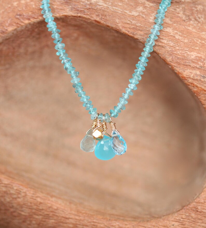 May include: A delicate necklace made with light blue gemstone beads and a charm of three smaller blue gemstones. The necklace is displayed on a wooden surface.