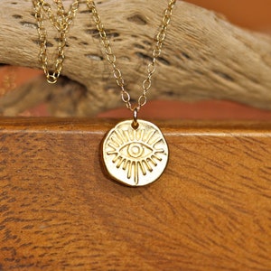 May include: Gold necklace with a round pendant featuring a stylized eye design. The pendant has a textured surface and a raised eye with a small circle in the center.