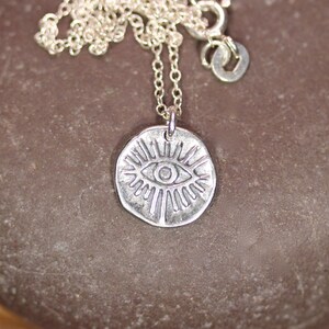 May include: Silver necklace with a round pendant featuring a stylized eye with radiating lines. The pendant is silver and has a textured finish.