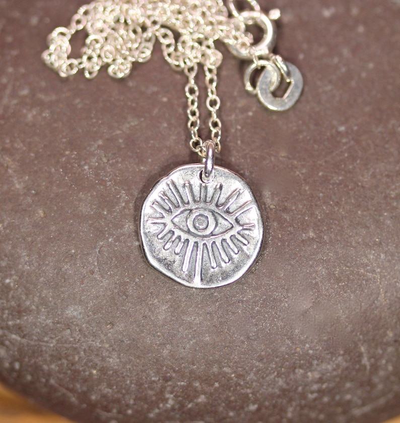 May include: Silver necklace with a round pendant featuring a stylized eye with radiating lines. The pendant is silver and has a textured finish.