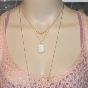 May include: A layered gold necklace with a white crystal pendant. The necklace is worn on a model wearing a pink crocheted top.