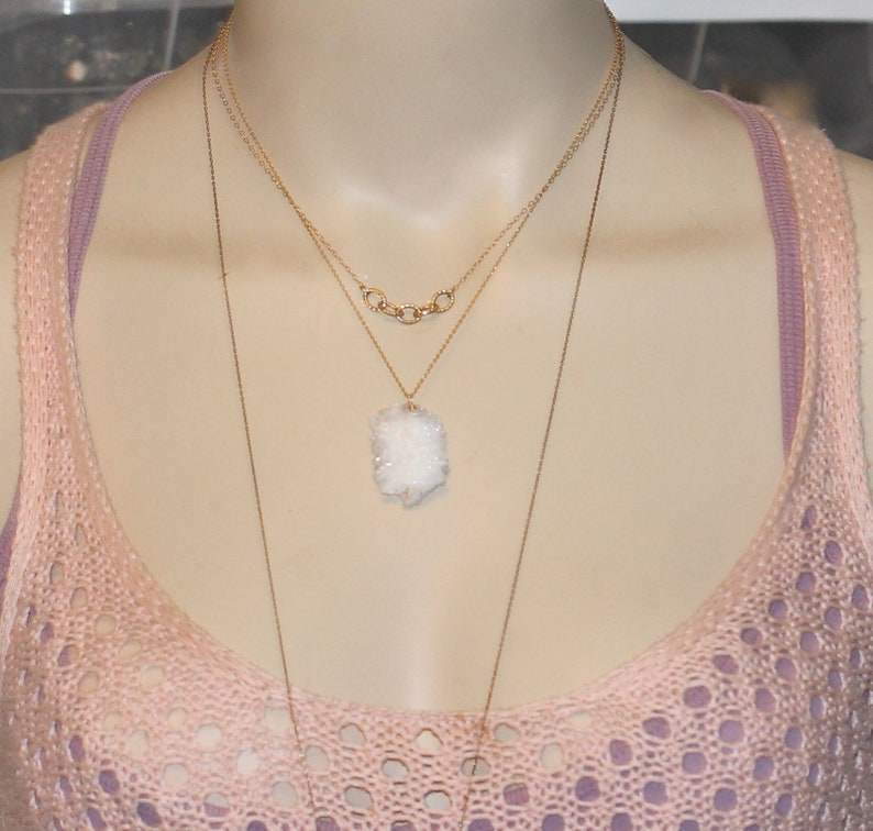 May include: A layered gold necklace with a white crystal pendant. The necklace is worn on a model wearing a pink crocheted top.