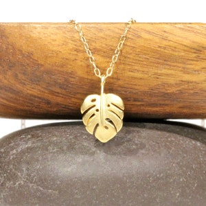 Peut inclure: Un collier en or avec un pendentif d&eacute;licat en forme de feuille de monstera d&eacute;coup&eacute;e. Le collier est pr&eacute;sent&eacute; sur une surface en bois et une pierre.
