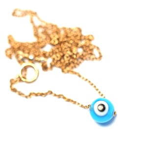 Puede incluir: Un collar de cadena dorada con un dije azul de ojo turco. El dije tiene un centro blanco con una pupila negra.