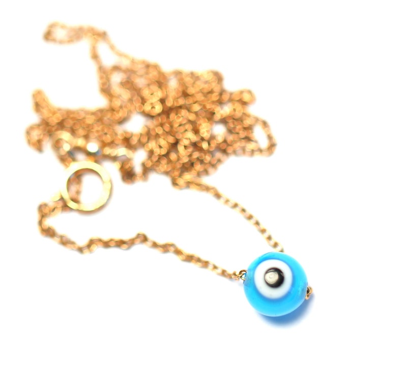 Puede incluir: Un collar de cadena dorada con un dije azul de ojo turco. El dije tiene un centro blanco con una pupila negra.
