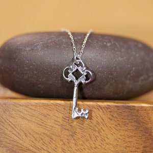 May include: A silver key pendant necklace with a detailed, ornate design. The key has a decorative, intricate top and a simple, straight shaft.