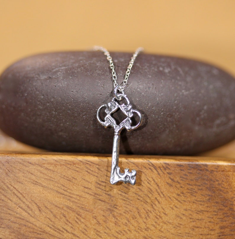 May include: A silver key pendant necklace with a detailed, ornate design. The key has a decorative, intricate top and a simple, straight shaft.