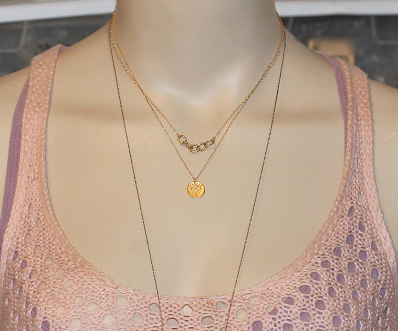 May include: A gold necklace with three chains, one with a small round pendant featuring a sun design. The necklace is worn on a mannequin with a pink crocheted top.