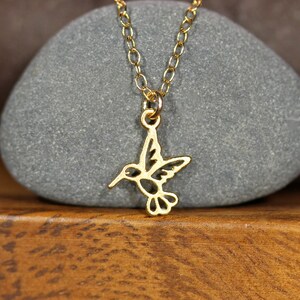 May include: A gold chain necklace with a delicate hummingbird pendant. The hummingbird is cut out of metal and has a detailed design.