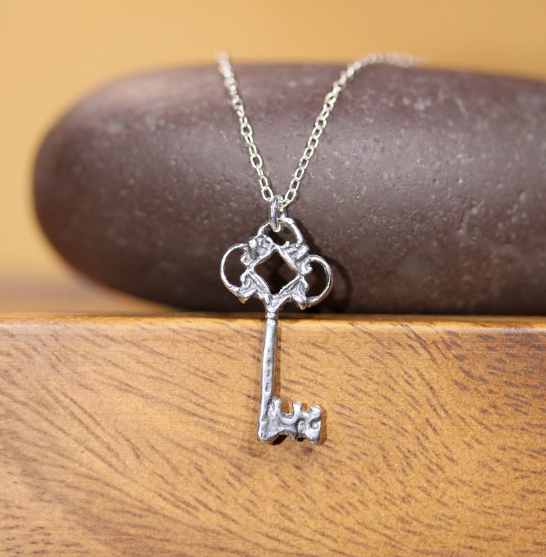 May include: A silver key pendant necklace with an ornate design. The key has a detailed, intricate design with a square in the center. The necklace is on a silver chain.