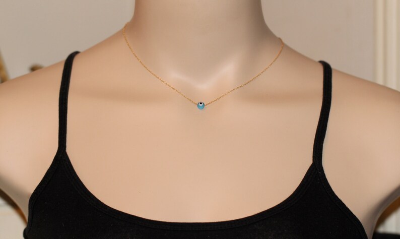 Puede incluir: Un collar de cadena dorada con un peque&ntilde;o dije azul de ojo turco. El collar est&aacute; en un maniqu&iacute; que lleva un top negro sin mangas.