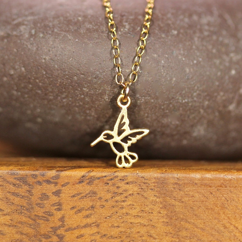 May include: A gold chain necklace with a delicate hummingbird pendant. The hummingbird is cut out of metal and has a detailed design.
