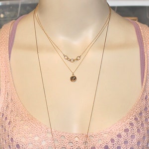 May include: Three gold necklaces layered on a mannequin. The top necklace has a series of connected circles. The middle necklace has a small round pendant. The bottom necklace is a long chain.