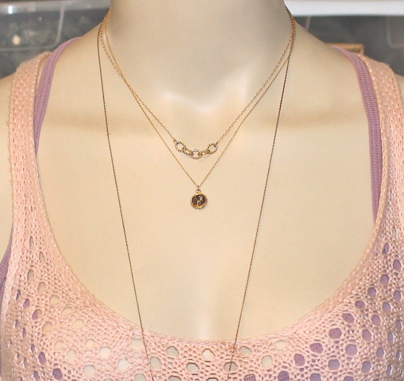 May include: Three gold necklaces layered on a mannequin. The top necklace has a series of connected circles. The middle necklace has a small round pendant. The bottom necklace is a long chain.