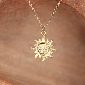 Pode incluir: Um colar de ouro com um pingente de sol. O sol tem um rosto com um sorriso e os olhos fechados.