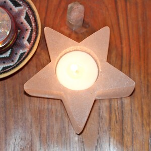 May include: A pink star-shaped candle holder with a round opening for a tea light candle. The candle holder is made of a textured material and is lit with a warm glow.