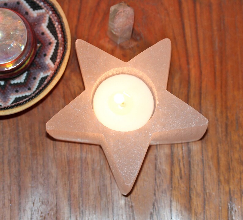 May include: A pink star-shaped candle holder with a round opening for a tea light candle. The candle holder is made of a textured material and is lit with a warm glow.