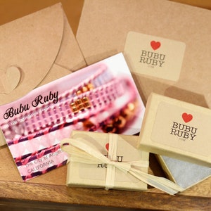 May include: A small brown box with a white label that says "Bubu Ruby" and a red heart. The box is wrapped in brown paper and tied with a string. The box is sitting on a wooden surface.