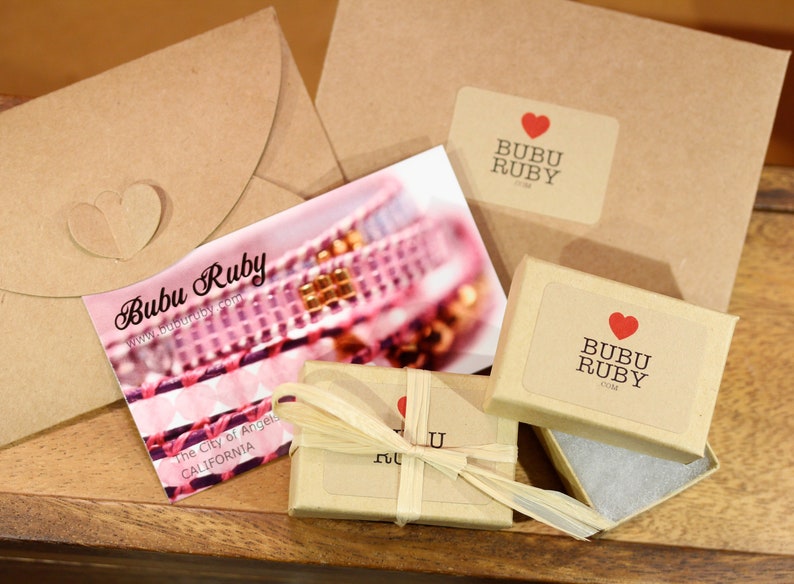 May include: A small brown box with a white label that says "Bubu Ruby" and a red heart. The box is wrapped in brown paper and tied with a string. The box is sitting on a wooden surface.