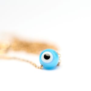 Puede incluir: Un collar de cadena dorada con un dije azul de ojo turco. El dije tiene un centro blanco y una pupila negra.