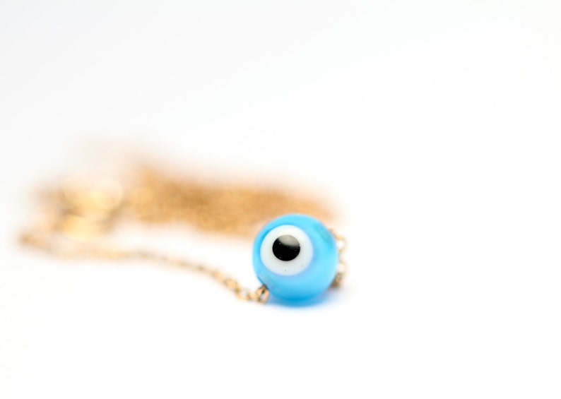 Puede incluir: Un collar de cadena dorada con un dije azul de ojo turco. El dije tiene un centro blanco y una pupila negra.