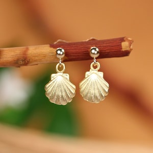 May include: A pair of gold-tone earrings with a seashell design. The earrings are dangling and have a small ball at the top.