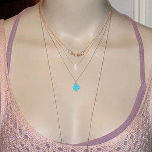 May include: A layered gold necklace with a turquoise pendant and a small white pendant. The necklace is worn on a mannequin with a pink tank top.