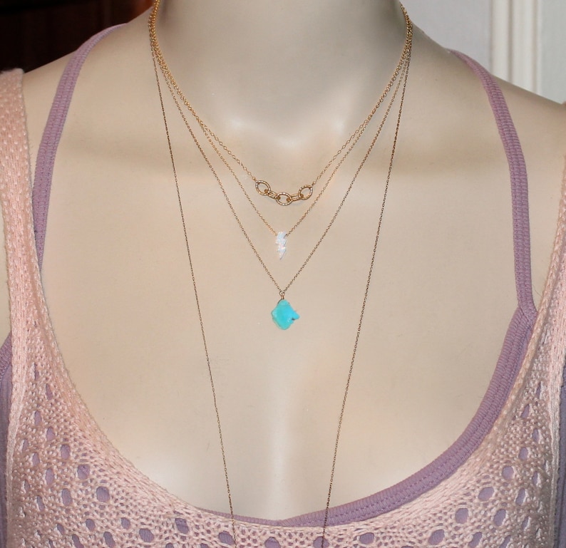 May include: A layered gold necklace with a turquoise pendant and a small white pendant. The necklace is worn on a mannequin with a pink tank top.