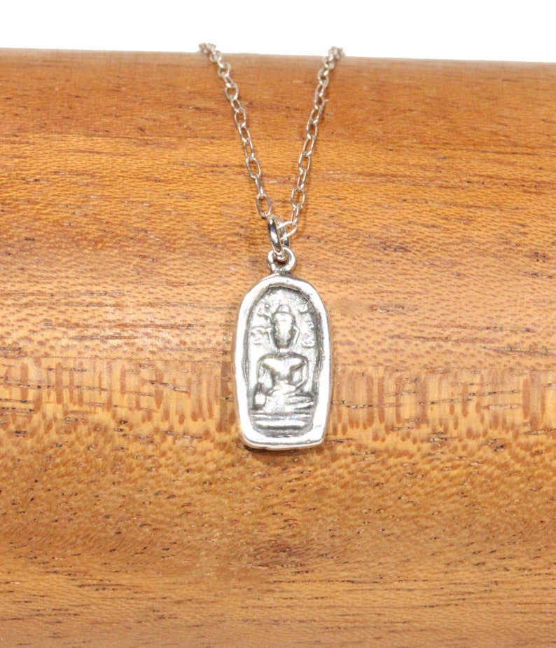 May include: A silver necklace with a pendant featuring a detailed image of a seated Buddha. The pendant is oval-shaped and has a textured surface.