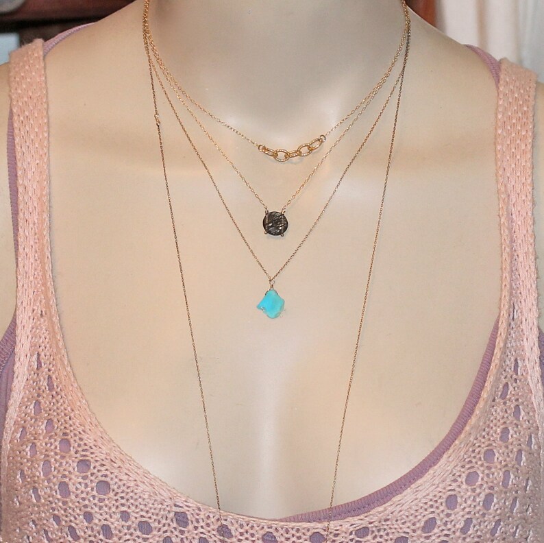 May include: A layered gold necklace with three pendants. The top pendant is a small, dark gray circle. The middle pendant is a larger, light blue, irregular shape. The bottom pendant is a small, light blue, irregular shape.