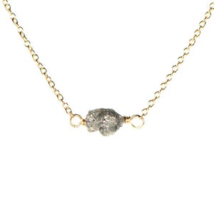 May include: A delicate gold chain necklace featuring a raw, grey diamond pendant. The diamond is irregularly shaped and has a rough, natural texture.