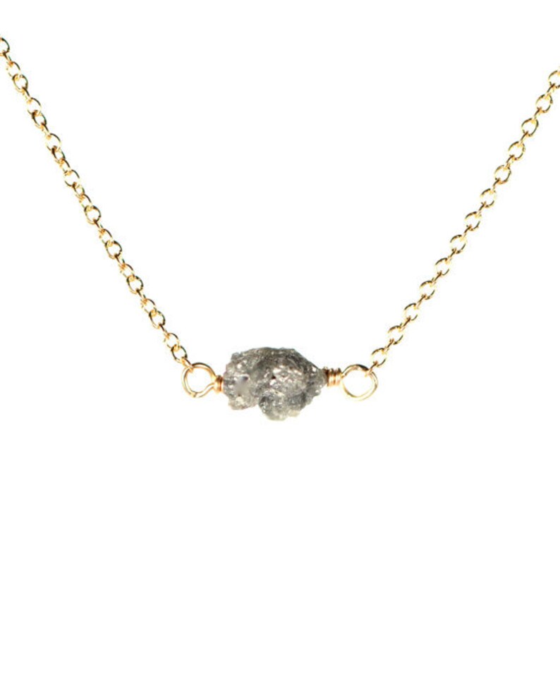 May include: A delicate gold chain necklace featuring a raw, grey diamond pendant. The diamond is irregularly shaped and has a rough, natural texture.