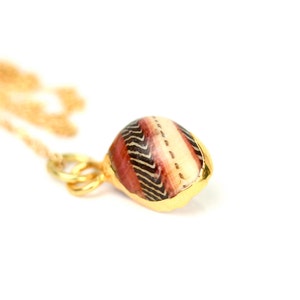 Può includere: Una collana con catena d'oro con un ciondolo fatto di una conchiglia con un motivo geometrico rosso, bianco e nero.