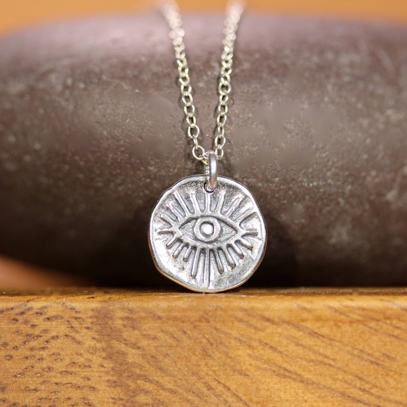 May include: Silver necklace with a circular pendant featuring a stylized eye design. The eye is surrounded by radiating lines.
