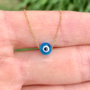 Puede incluir: Un collar de cadena dorada con un dije azul de ojo turco. El dije tiene un centro blanco y una pupila negra.