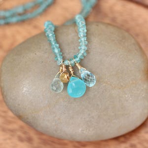 May include: A delicate necklace made with light blue gemstone beads and a charm of three teardrop-shaped gemstones in shades of blue and gold.