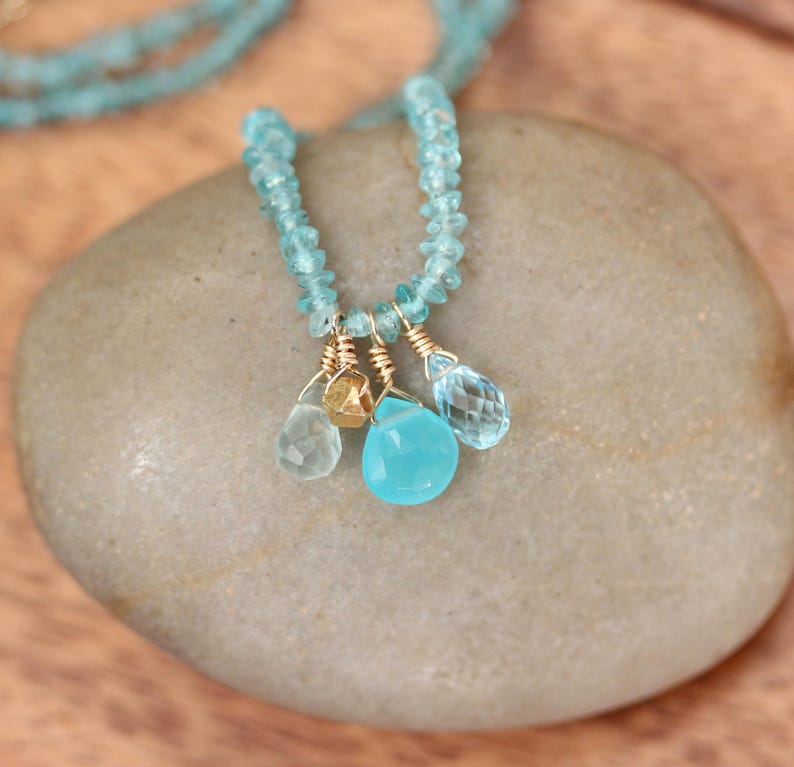May include: A delicate necklace made with light blue gemstone beads and a charm of three teardrop-shaped gemstones in shades of blue and gold.