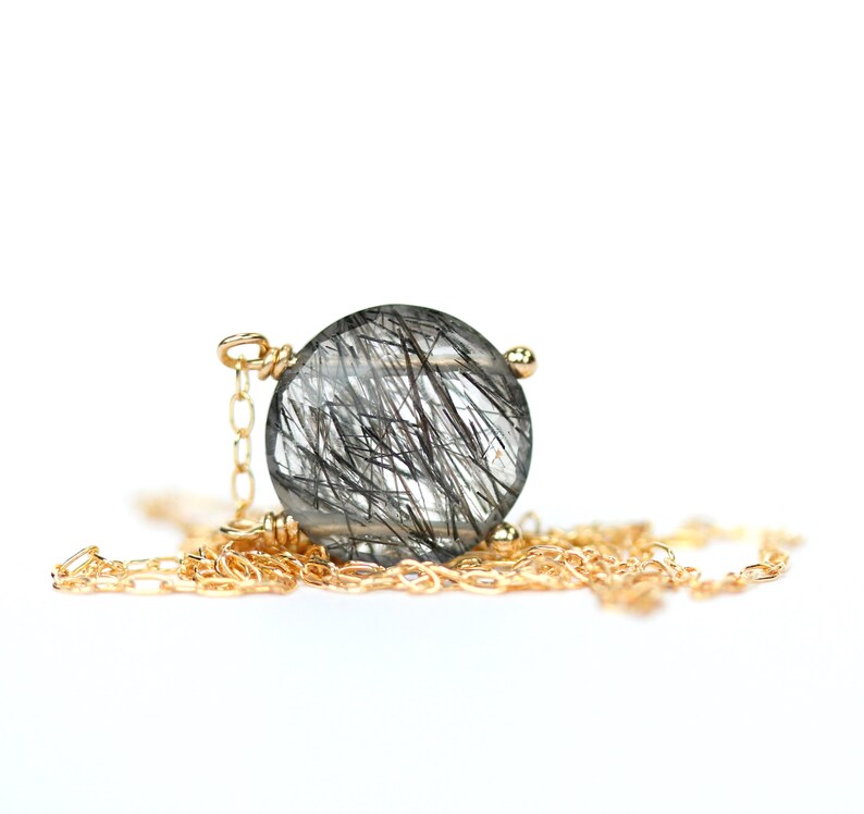 May include: A gold chain necklace with a round, clear quartz pendant featuring black inclusions. The pendant is set in a gold bezel.