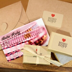 May include: A small brown box with a white label that says "Bubu Ruby" and a red heart. The box is wrapped in brown paper and tied with a string. The box is sitting on a wooden surface.