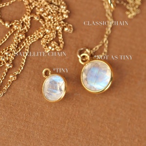 May include: Two gold necklaces with moonstone pendants. The necklace on the left has a satellite chain and a smaller pendant labeled "*TINY". The necklace on the right has a classic chain and a larger pendant labeled "*NOT AS TINY".